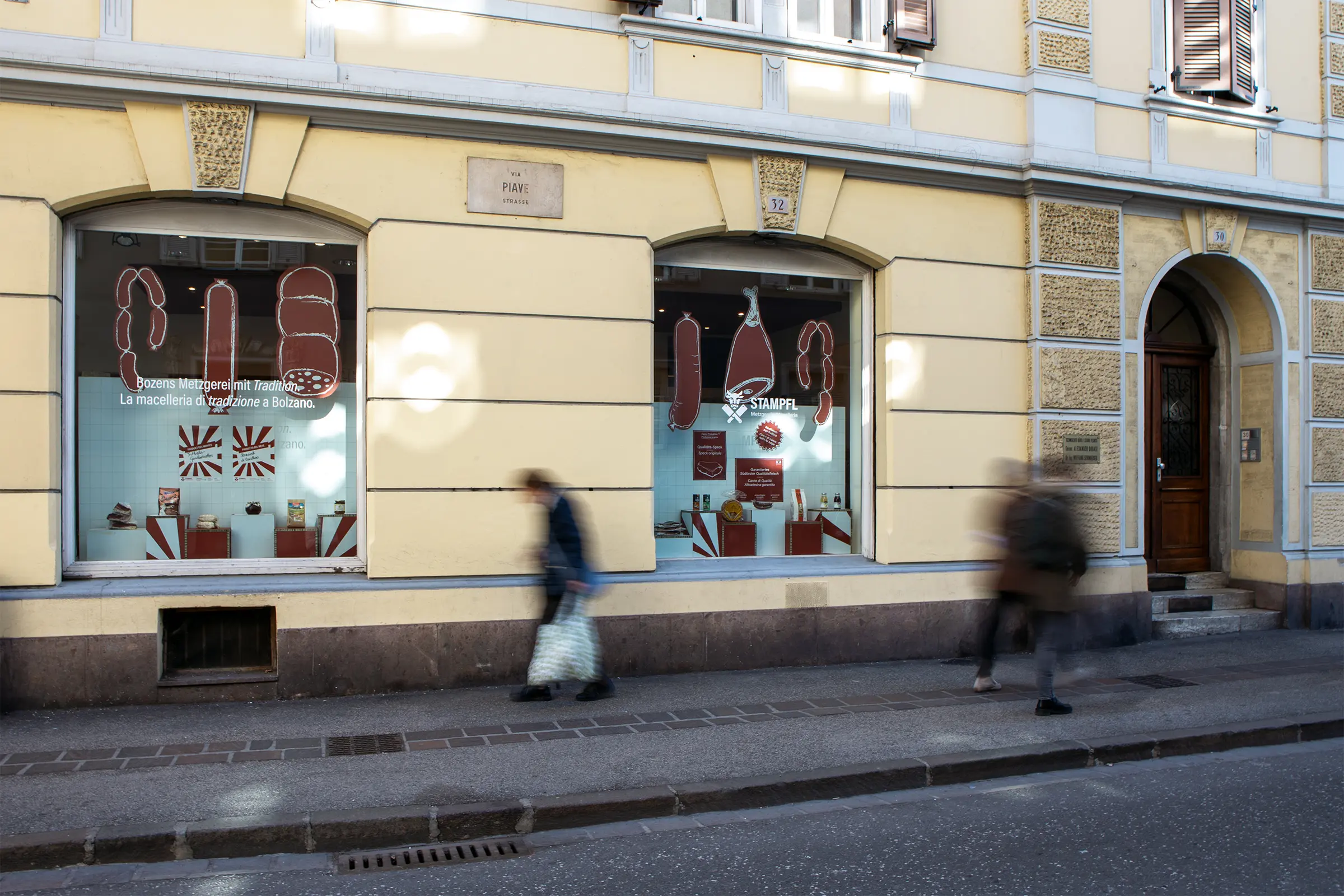 Zwei Schaufenster der Metzgerei Stampfl mit Auslage, während drei Passant:innen auf dem Gehweg vorbeigehen.