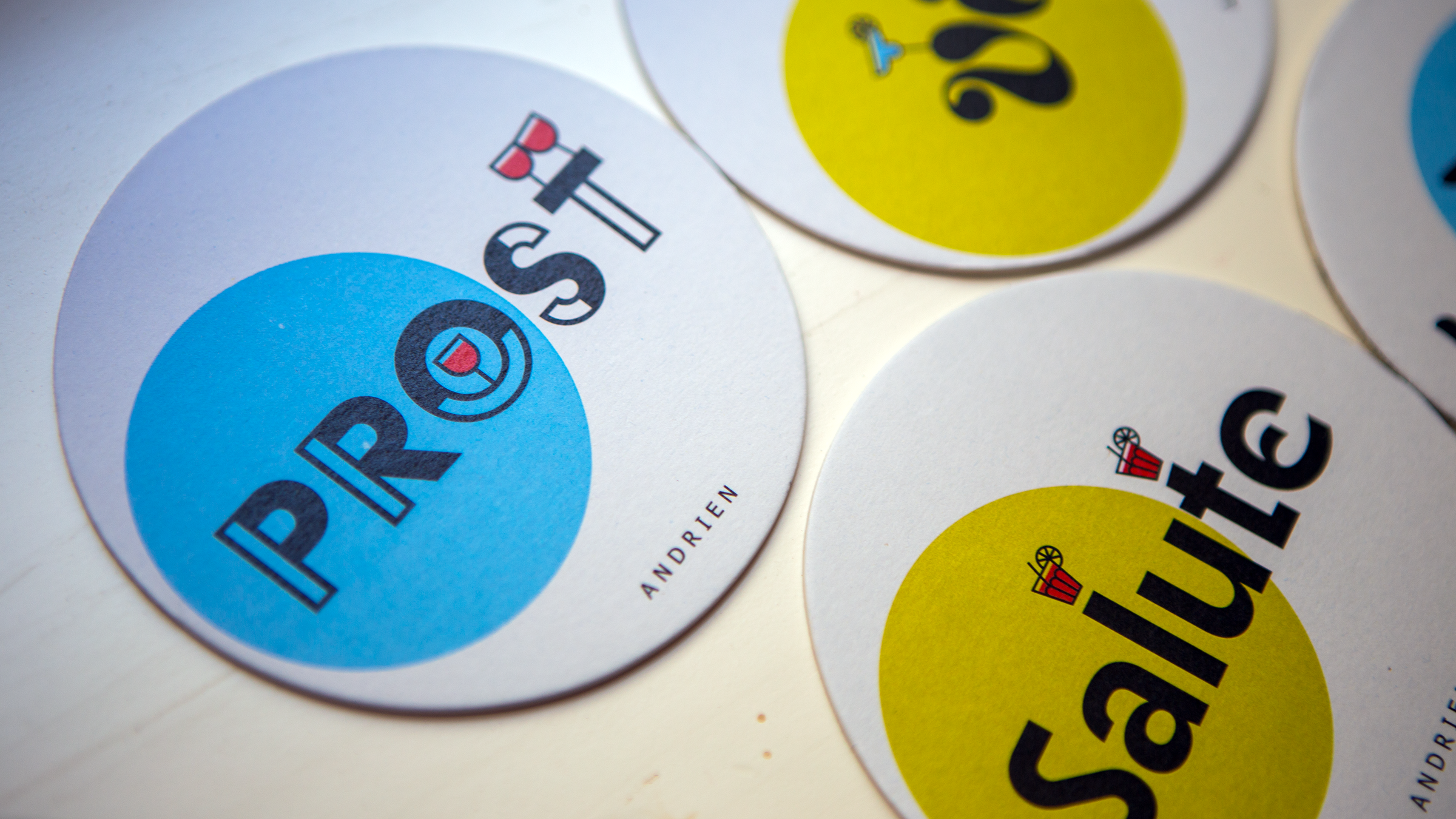 Drei Bierdeckel mit Südtiroler Trinksprüchen, blau-gelben Farbelementen und kleinen Getränke-Illustrationen.