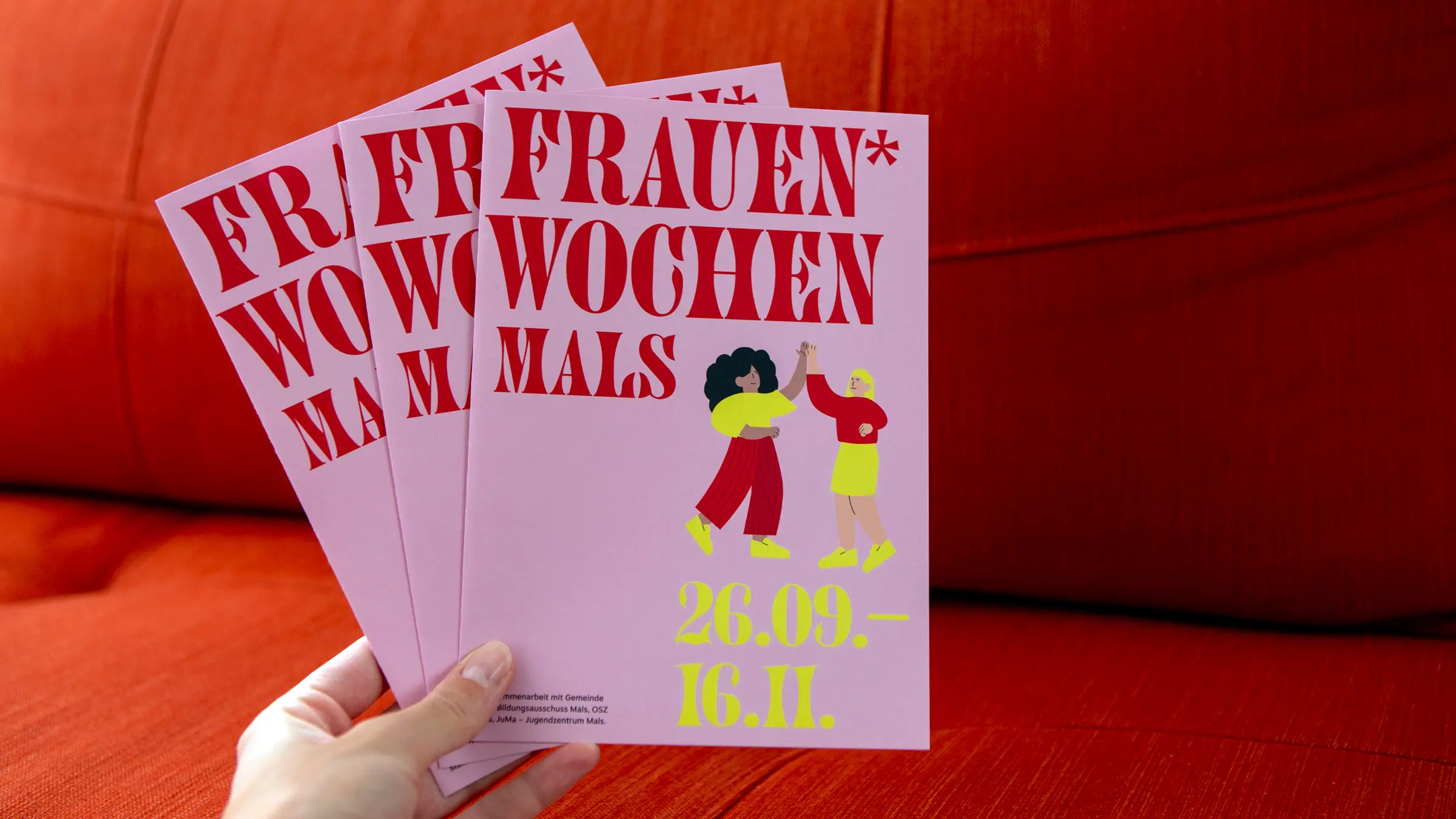 Drei aufgefächerte Flyer für die Frauenwochen in Mals mit roten und gelben Schriftzügen sowie Frauen-Illustrationen auf rosa Hintergrund.