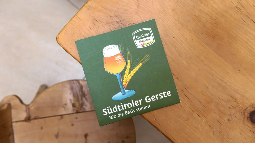 Auf der Kante eines rustikalen Tisches liegt eine Broschüre für Südtiroler Gerste mit grünem Hintergrund und der Illustration eines Bierglases und zwei Kornähren.