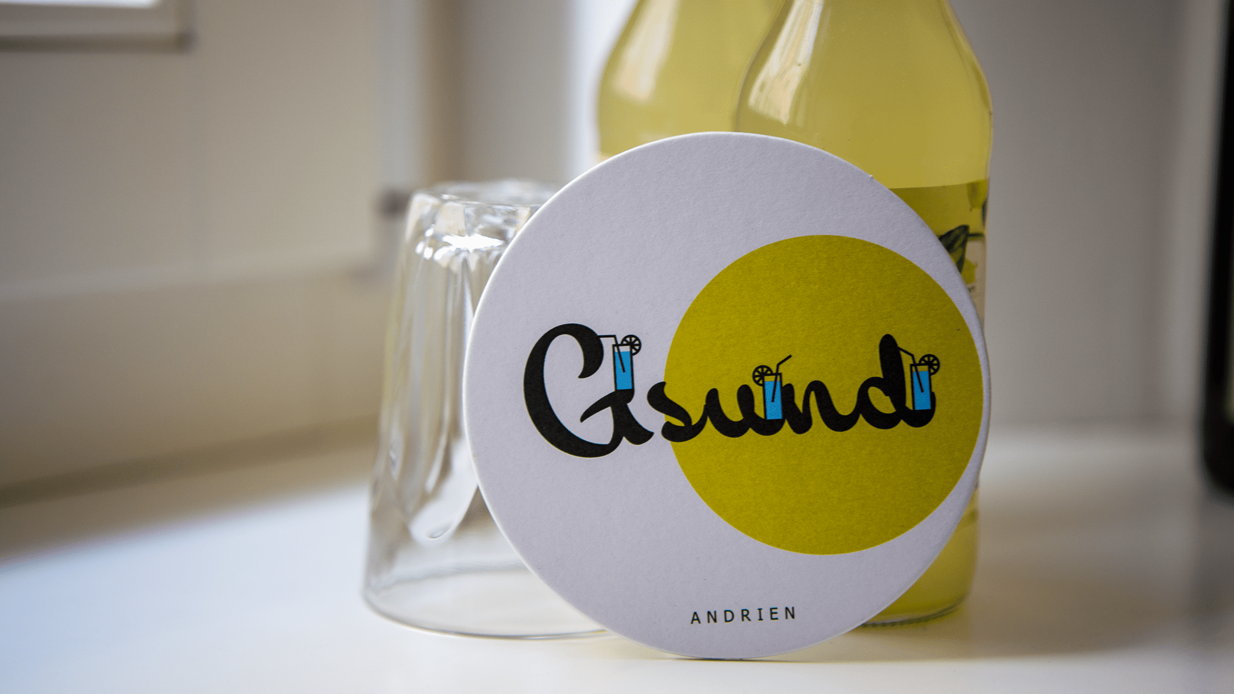 An einer Limonadenflasche und einem Trinkglas lehnt ein Bierdeckel des Hofschanks Andrien mit Südtiroler Trinkspruch, gelbem Farbelement und Cocktailglas-Illustrationen.