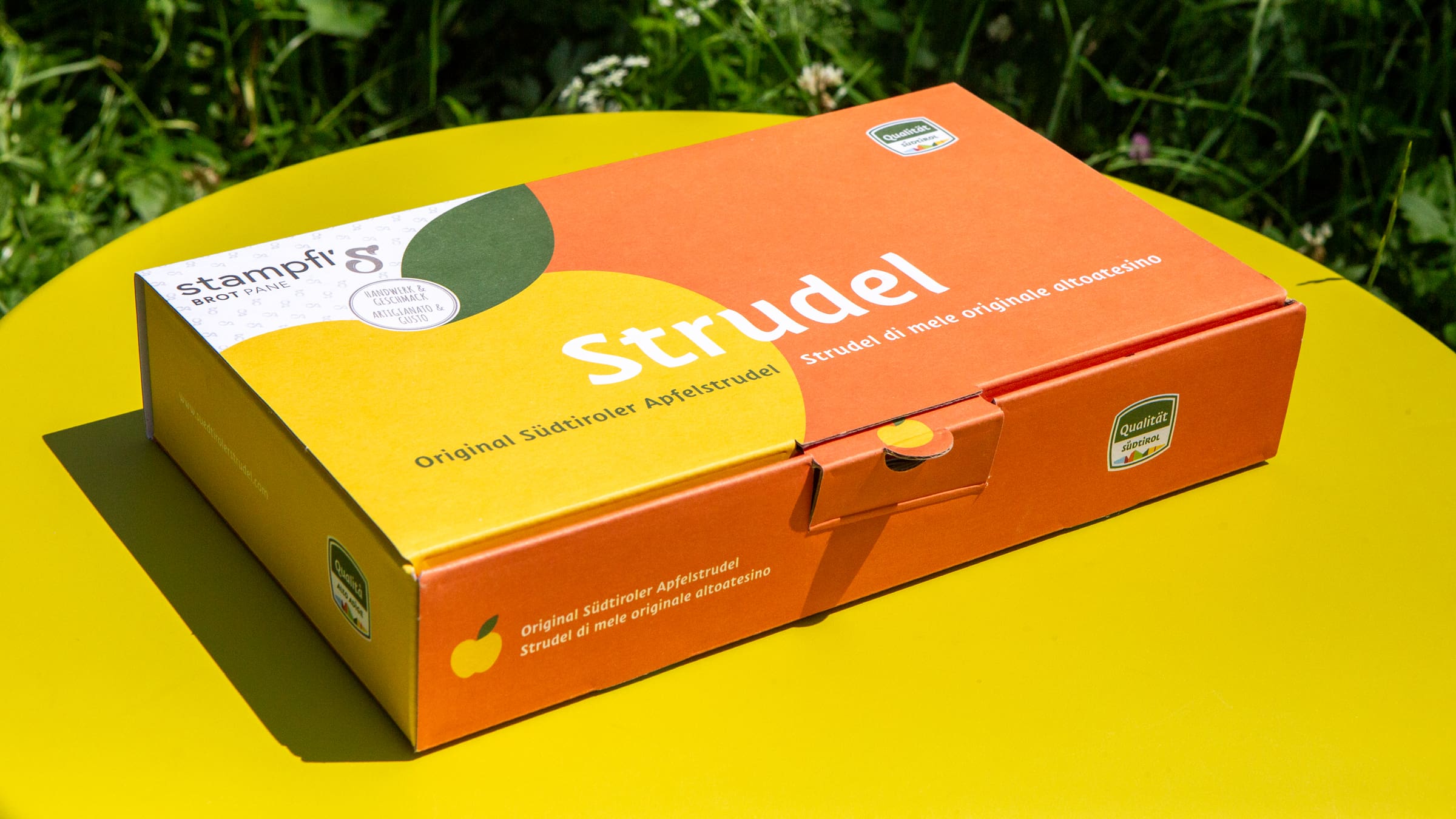 Kartonverpackung für Südtiroler Apfelstrudel mit Apfel-Illustrationen auf orangem Hintergrund.