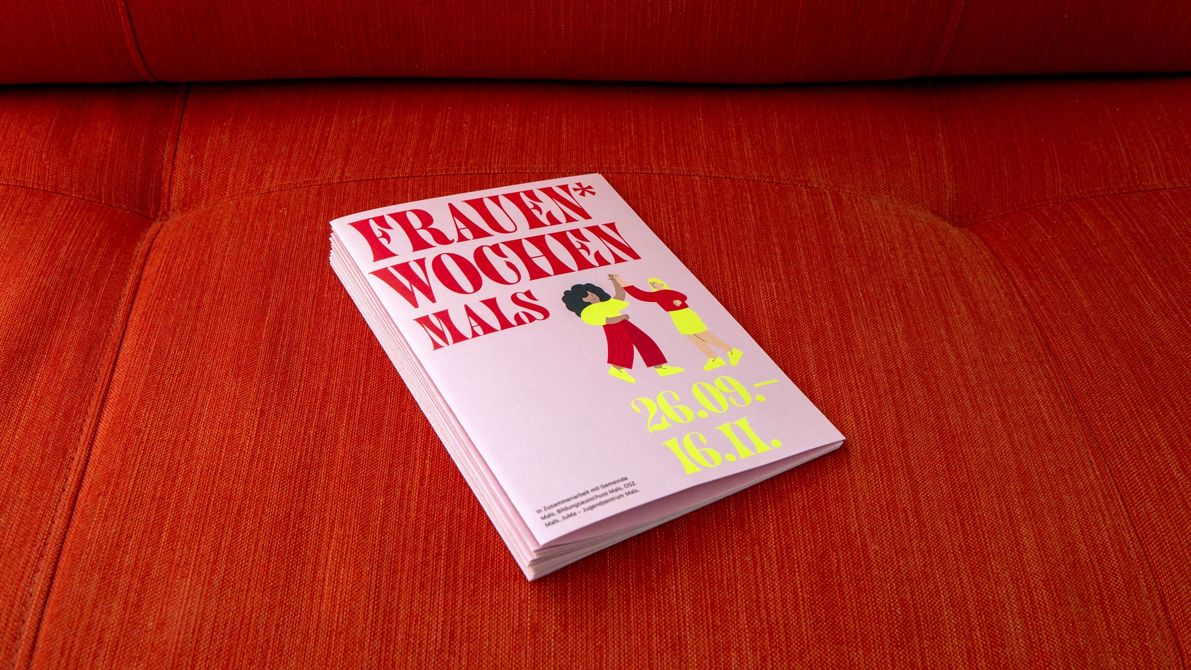 Stapel mit Flyern für die Frauenwochen in Mals mit roten und gelben Schriftzügen sowie Frauen-Illustrationen auf rosa Hintergrund.