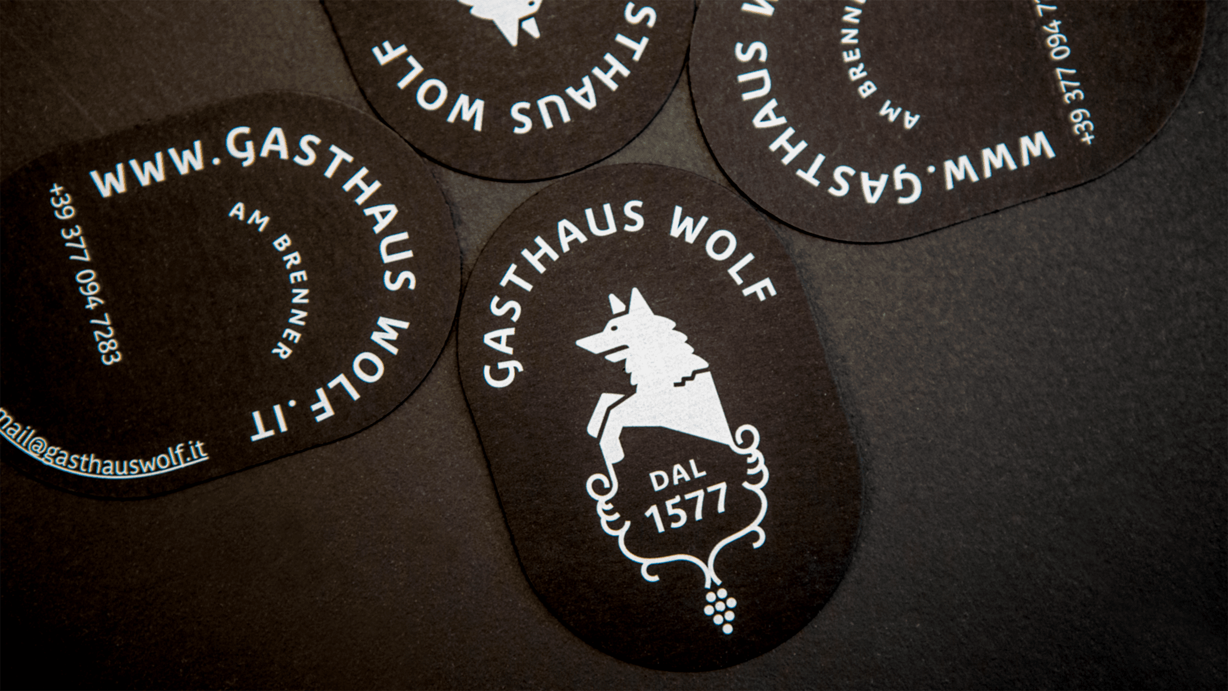 Visitenkarten des Gasthauses Wolf mit weißem Logo auf schwarzem Papier in ovaler Form.